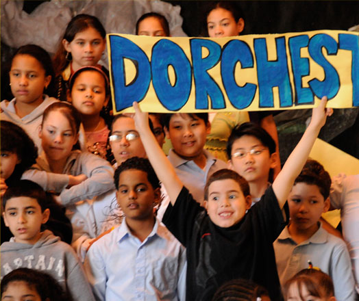 A student in a group with others holds up a sign that reads "Dorchester"