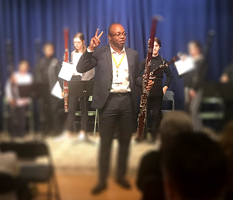 A man holds a bassoon as he talks to an audience